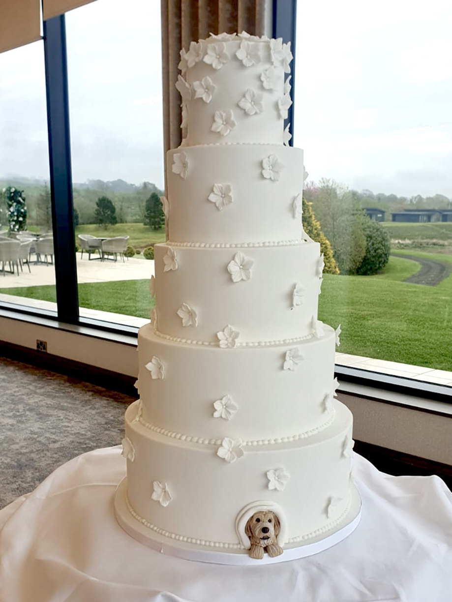 White five-tier cake with icing flowers and dog made of icing peering out from bottom tier